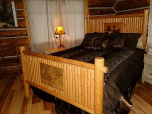 Una cama con estructura de madera y un edredón oscuro, una mesita de noche con una lámpara, y una ventana con cortinas.