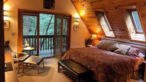 A cozy bedroom with a large bed, a bench, a desk with a chair, and a balcony with a view of trees.