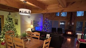 A cozy cabin interior with a dining area, two Christmas trees, a fireplace, and a large window.