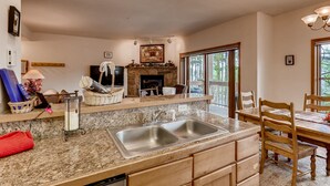 A kitchen with granite countertops, a double sink, a stone fireplace, and a dining area with wooden chairs.
