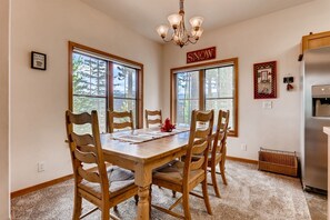 Un coin repas avec une table et des chaises en bois, de grandes fenêtres, un lustre et un réfrigérateur.