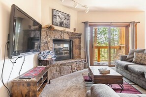 A living room with a stone fireplace, a flat-screen TV, a sofa, and a large window with a view of trees. - Breckenridge Luxury Condo, Easy Access to Ski Lifts! (Breckenridge)