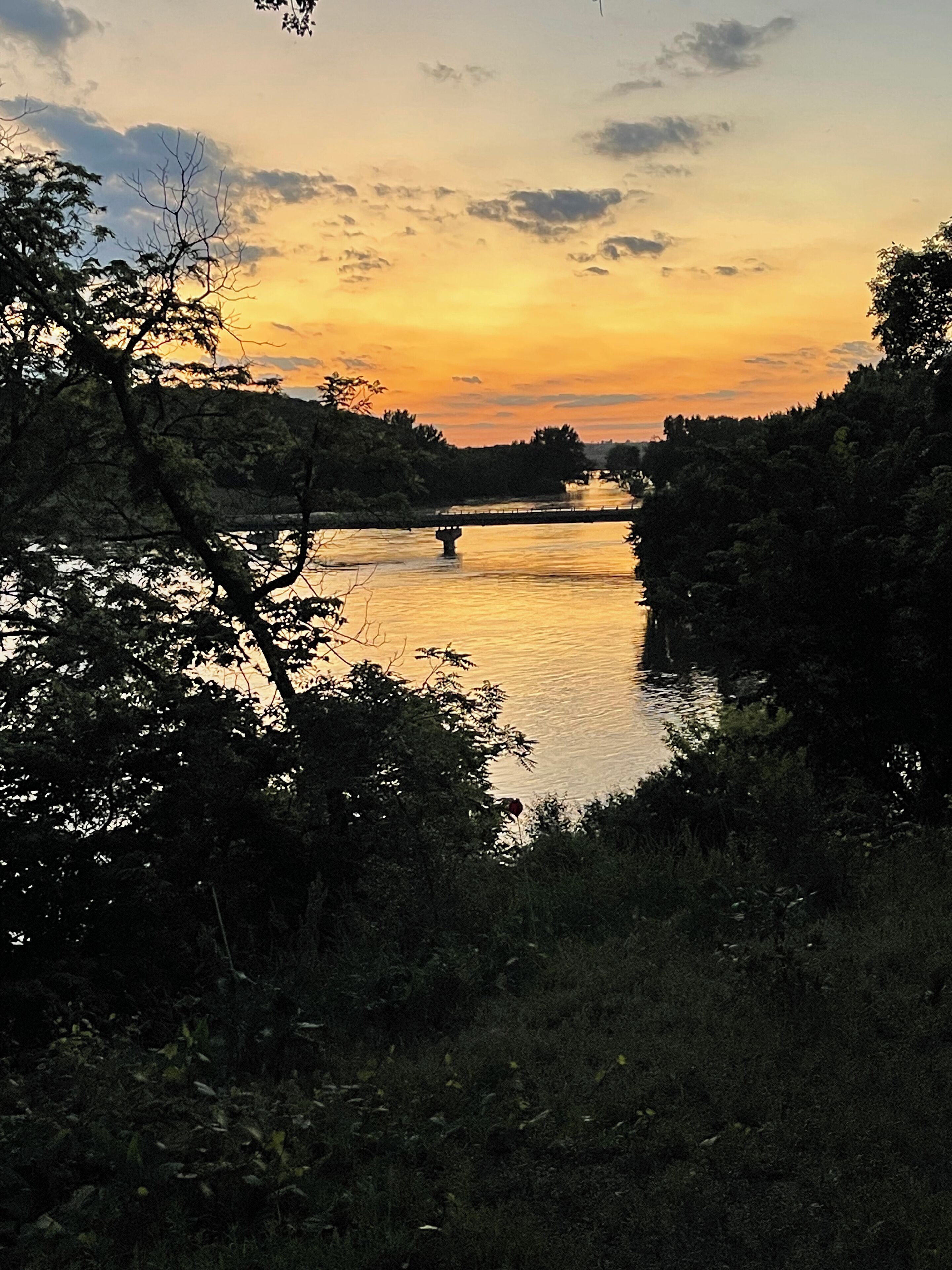 Une vue sur un coucher de soleil au-dessus d’une rivière, avec un pont, le tout entouré d’une végétation luxuriante.