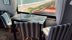Ein Zimmer mit Blick auf den Strand, ein Tisch mit einer Vase, gestreifte Stühle und ein Fenster mit Vorhängen.