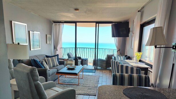 Ein Wohnzimmer mit Sofa, Sessel, Couchtisch und Fernseher. Ein Balkon bietet Blick aufs Meer.
