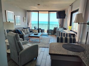Un salon comprenant un canapé, un fauteuil, une table basse et un téléviseur. Il y a également un balcon avec vue sur la mer.