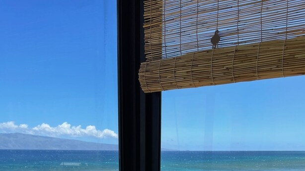 Habitación con vista al océano a través de un ventanal grande, una persiana de bambú y una barandilla de metal.