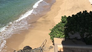 Una playa con agua cristalina y orilla de arena, un letrero de advertencia y edificios al fondo.