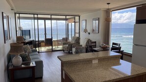 Amplia sala de estar con un gran ventanal corredizo que da a un balcón con vistas al océano. Cuenta con una zona de cocina con encimeras de granito y una mesa de comedor.