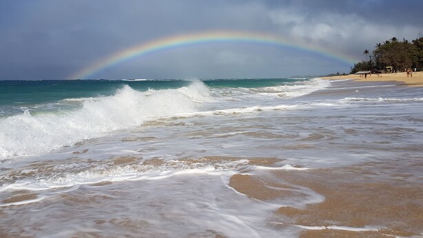 Ein Strand mit Wellen, einem Regenbogen und blauem Himmel.