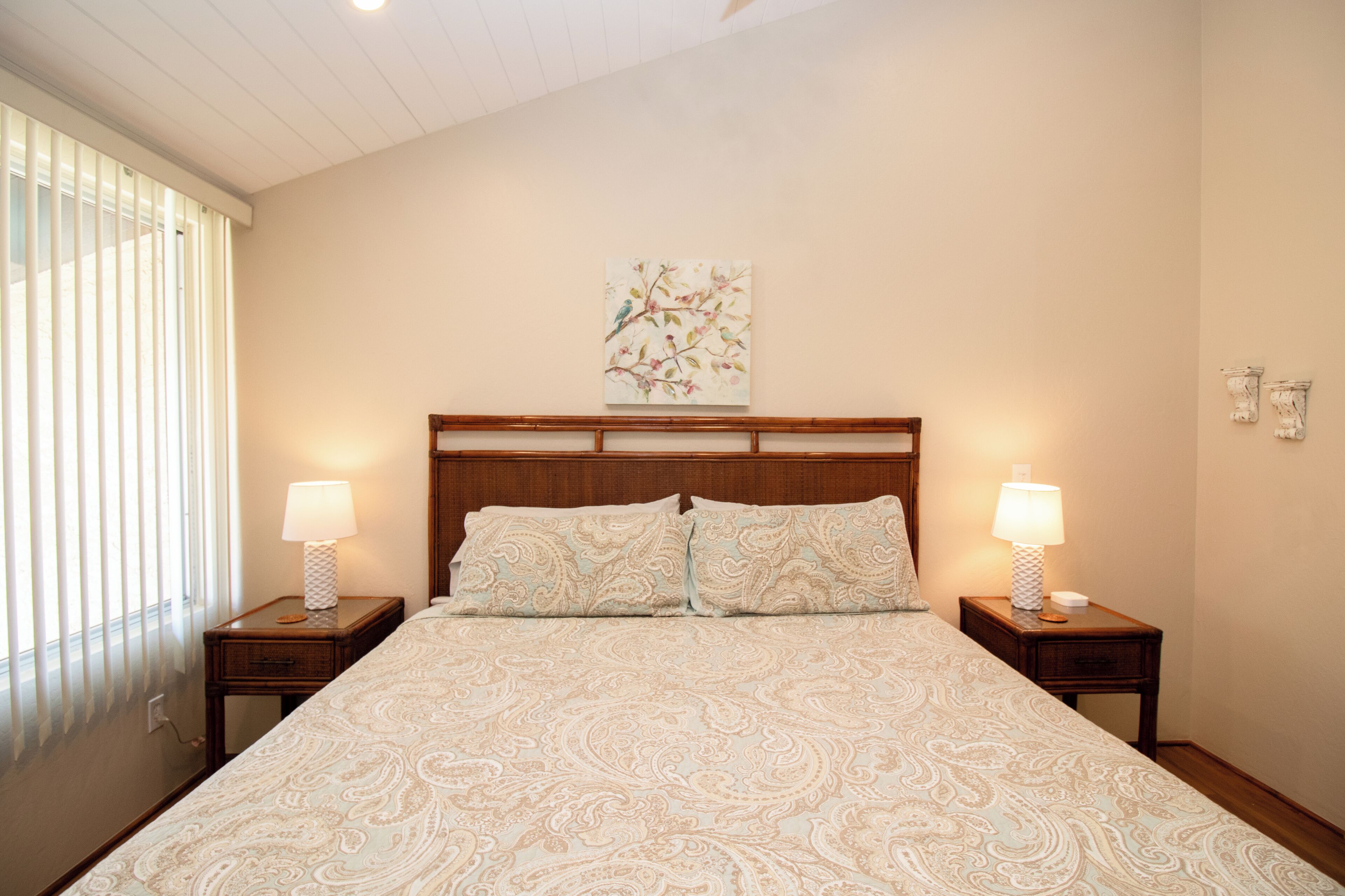 Une chambre à coucher avec un grand lit, deux tables de chevet avec des lampes, une tête de lit en bois et une œuvre d’art aux motifs floraux accrochée au mur.