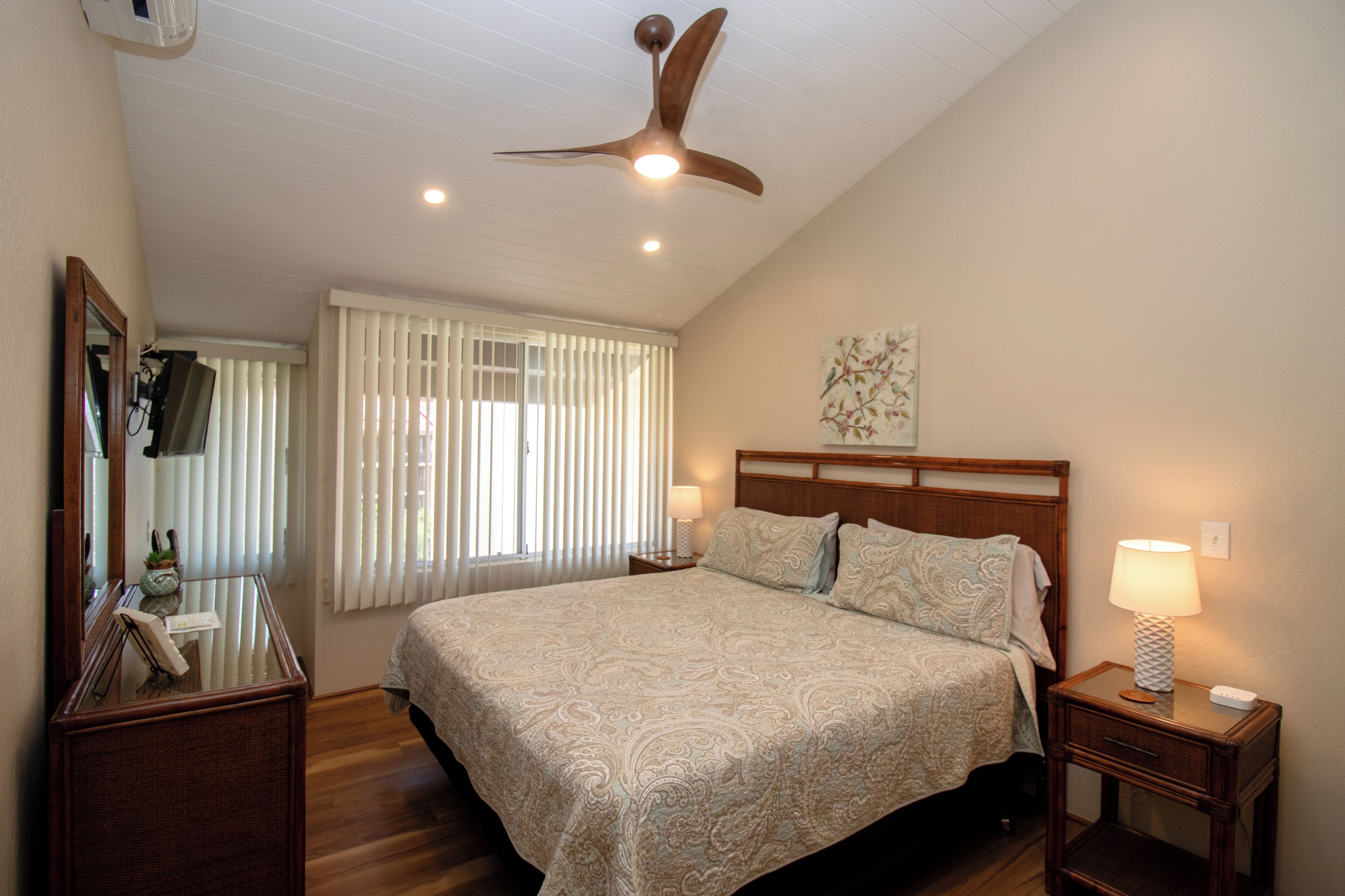 Une chambre avec un grand lit, un ventilateur de plafond, une table de chevet avec une lampe, une commode, un miroir et une fenêtre avec des stores.