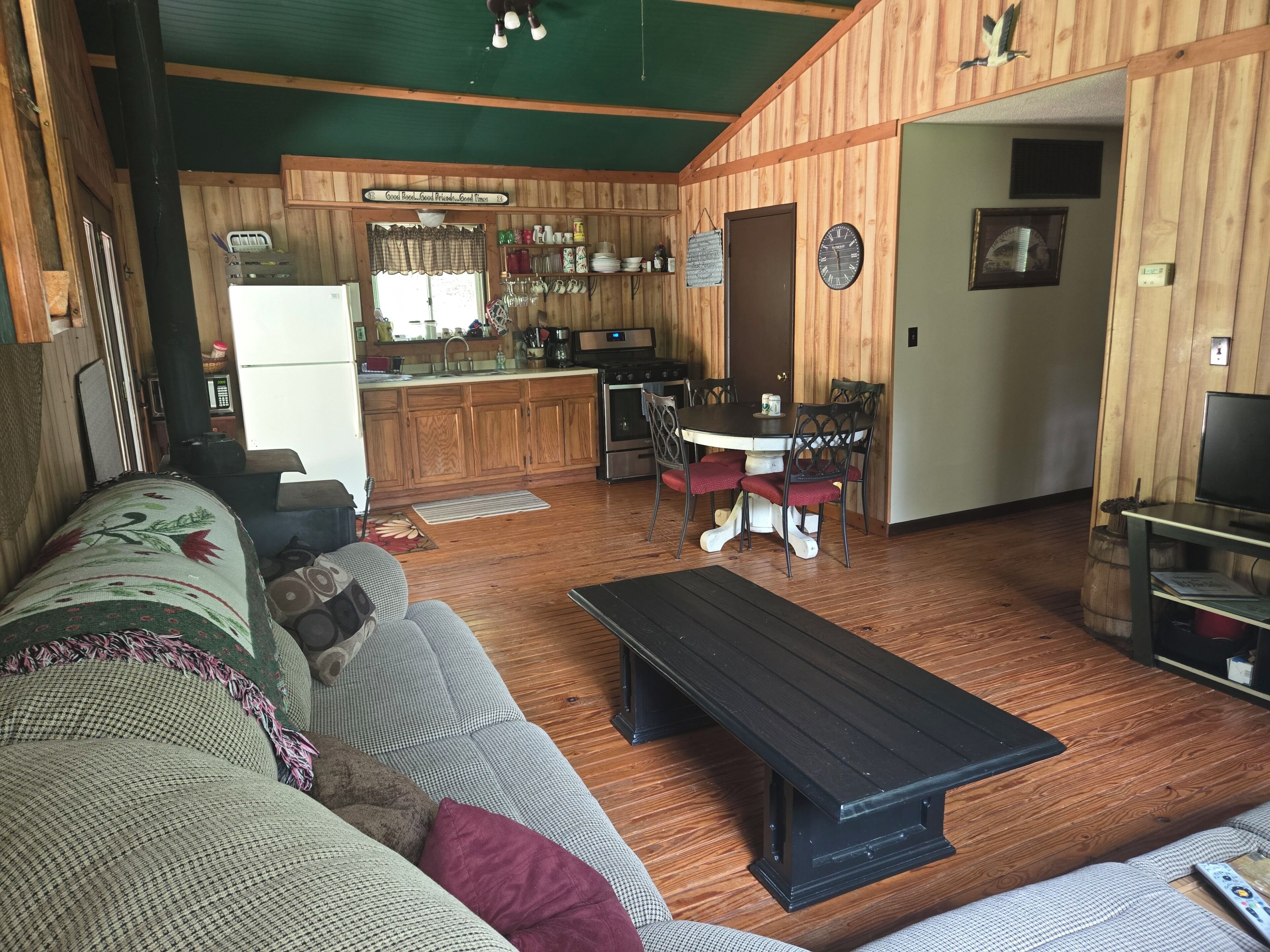A cozy living room with a sofa, a coffee table, and a dining area with a table and chairs. There is a kitchen area with wooden cabinets and a refrigerator.