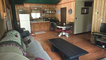 A cozy living room with a sofa, a coffee table, and a dining area with a table and chairs. There is a kitchen area with wooden cabinets and a refrigerator.