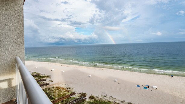 A beach view from a balcony with a railing, a sandy beach, and a rainbow in the distance. - Free parking til Nov. 15! 162 reviews, wrap around balcony, stay 7 pay for 6! OB (Orange Beach)