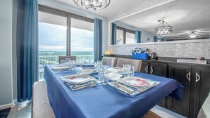 A dining area with a table set for four, large windows with curtains, a view of the city, and a kitchen area with dark cabinets and a backsplash. - Free parking til Nov. 15! 162 reviews, wrap around balcony, stay 7 pay for 6! OB (Orange Beach)