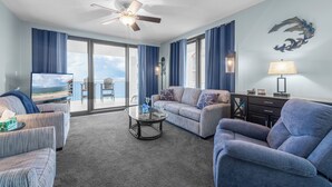 A spacious living room with a ceiling fan, a television, and a view of the beach through large windows. - Free parking til Nov. 15! 162 reviews, wrap around balcony, stay 7 pay for 6! OB (Orange Beach)