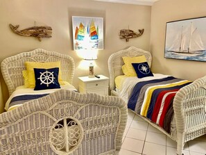 Two wicker beds with nautical-themed pillows, a nightstand with a lamp, and wall art including a sailboat and a colorful sailboat illustration. - Breathtaking Oceanfront Views at Brigadune – Luxury Condo on Quiet Shore Drive (Myrtle Beach)