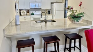 A modern kitchen with white cabinets, a stainless steel sink, a microwave, a stove, and a refrigerator. There are three wooden stools and a potted plant on the counter.