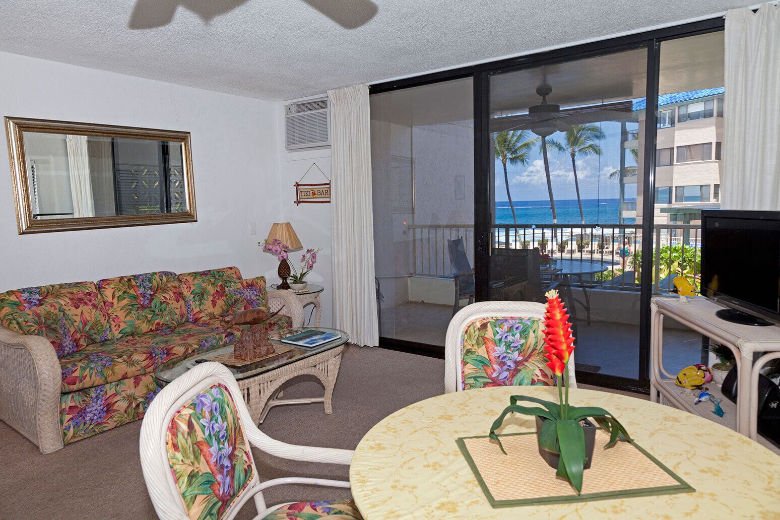 A living room with a sofa, a coffee table, and a TV. There is a balcony with a view of the ocean and palm trees.