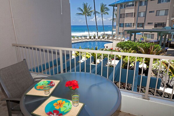 A balcony with a table set for two, overlooking a pool area with lounge chairs and palm trees.