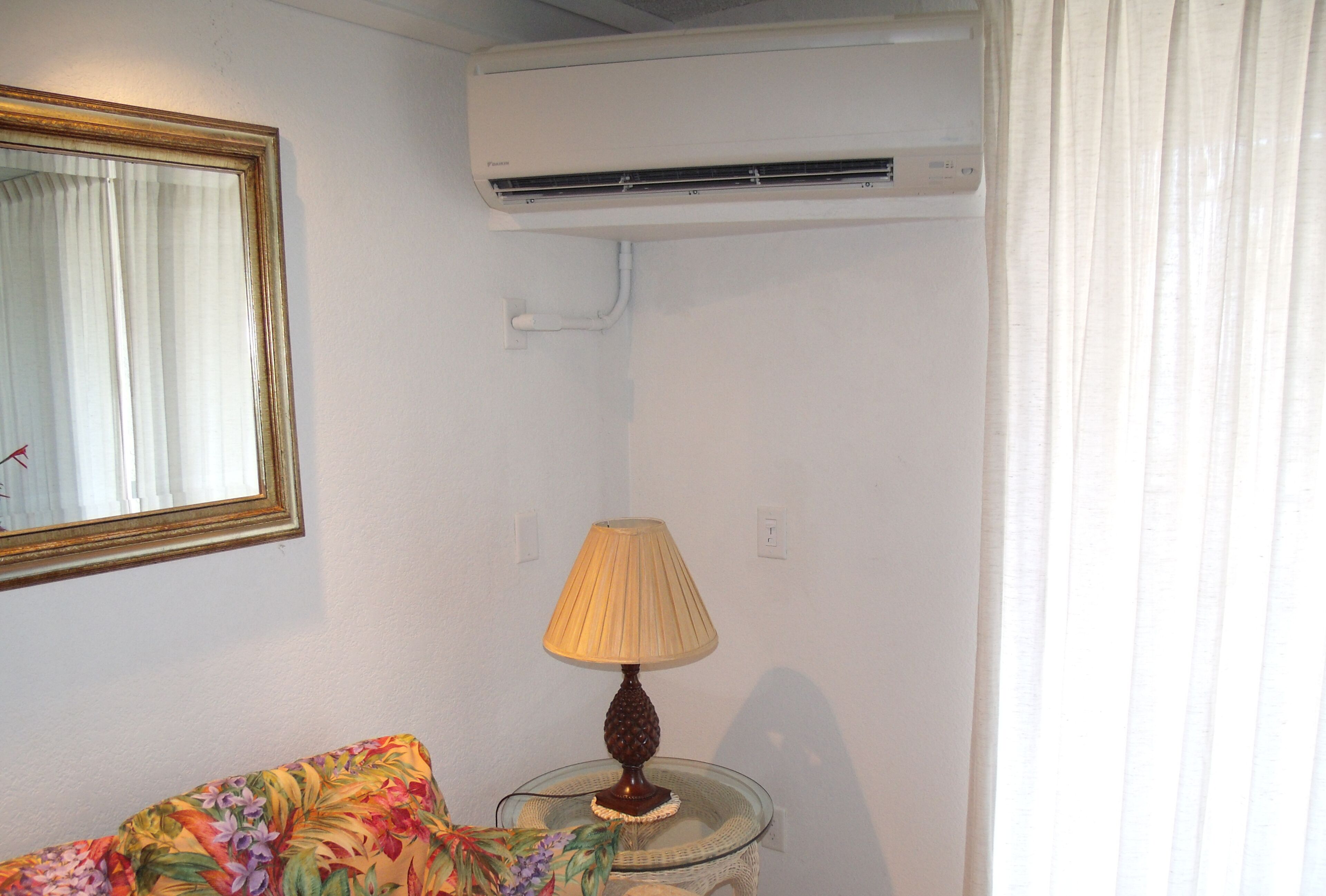 A room with a single bed, a bedside table with a lamp, an air conditioner, and a mirror on the wall.