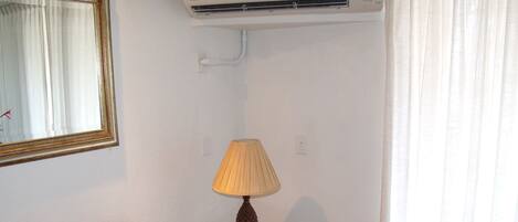 A room with a single bed, a bedside table with a lamp, an air conditioner, and a mirror on the wall.