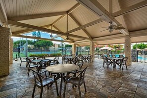 An outdoor patio area with round tables and chairs, a ceiling fan, and a view of a swimming pool and greenery. - Ocean, Beach, Pool - Just Steps Away - AC in both bedrooms! Beautiful Townhouse! (Kailua-Kona)