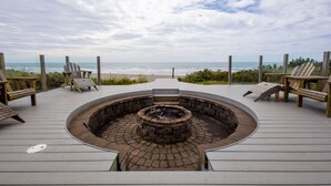 A circular fire pit surrounded by wooden deck chairs, overlooking a beach. - Ocean Front Home in the Heart of Newport! (Newport)
