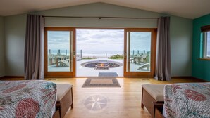 A bedroom with two beds, a large sliding glass door leading to a deck with a fire pit, and a view of the ocean. - Ocean Front Home in the Heart of Newport! (Newport)