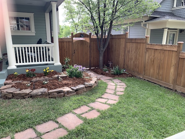 Patio trasero residencial con un sendero de piedra, una cerca de madera y diversas plantas.