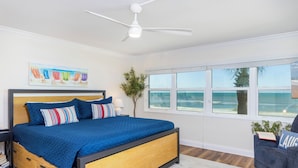 Un dormitorio con cama, ventilador de techo, ventana con vista a la playa y un cuadro en la pared.