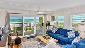 A living room with a view of the ocean, a ceiling fan, and a blue sofa.