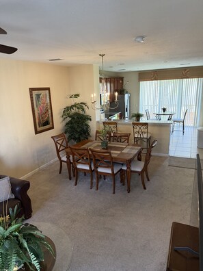 A dining area with a round table, chairs, and a chandelier. There is a kitchen in the background with cabinets and a refrigerator.