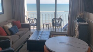 Un salon comprenant un canapé, un pouf et une table ronde, un balcon avec vue sur la mer, et un téléviseur fixé au mur.