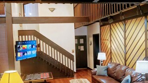 A living room with a brown leather couch, a coffee table, a TV mounted on the wall, and a staircase leading to an upper level.