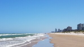 A beach with a sandy shore and waves, a clear sky, and buildings in the background.
