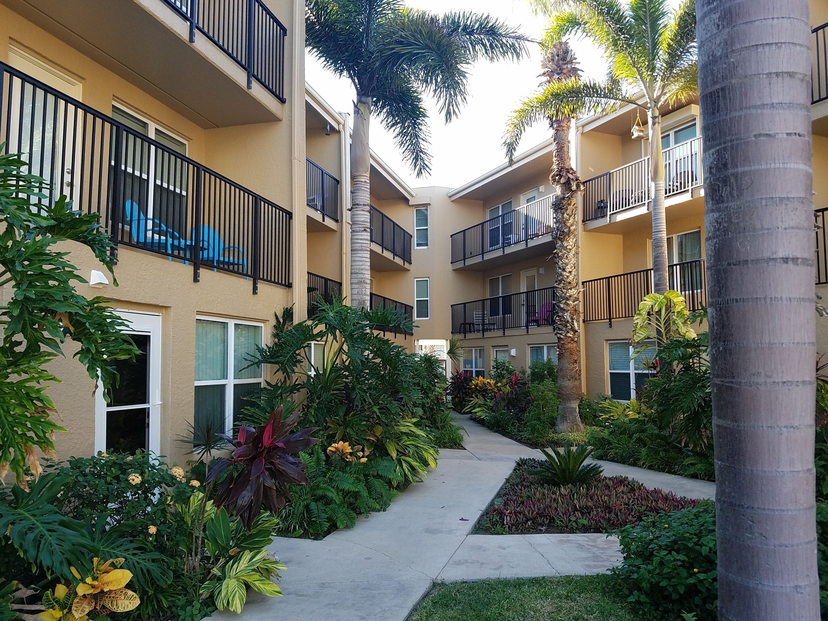 A multi-story residential building with balconies, a central pathway, and various tropical plants.