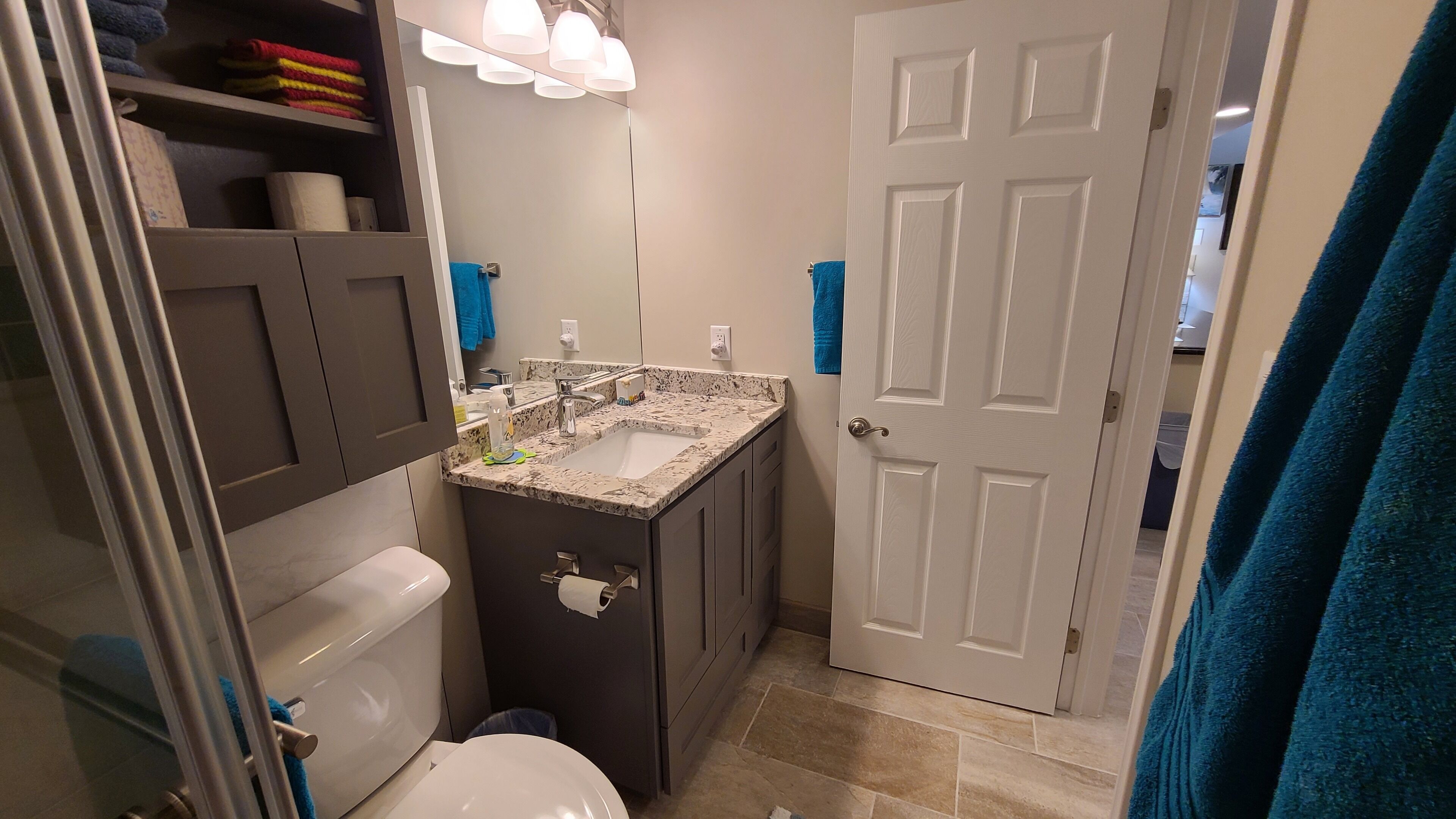 A bathroom with a white toilet, a sink with a marble countertop, and a mirror above the sink. There is a towel rack with blue towels and a door with a white doorbell.