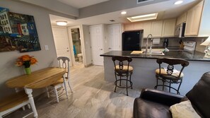A compact kitchen with a dining area, featuring a wooden table, two bar stools, and a refrigerator. - SURFSIDE 1  107 BEACH Condo 1 KING 2TVs Washerdyer HEATED POOL Hot tub (South Padre Island Texas)
