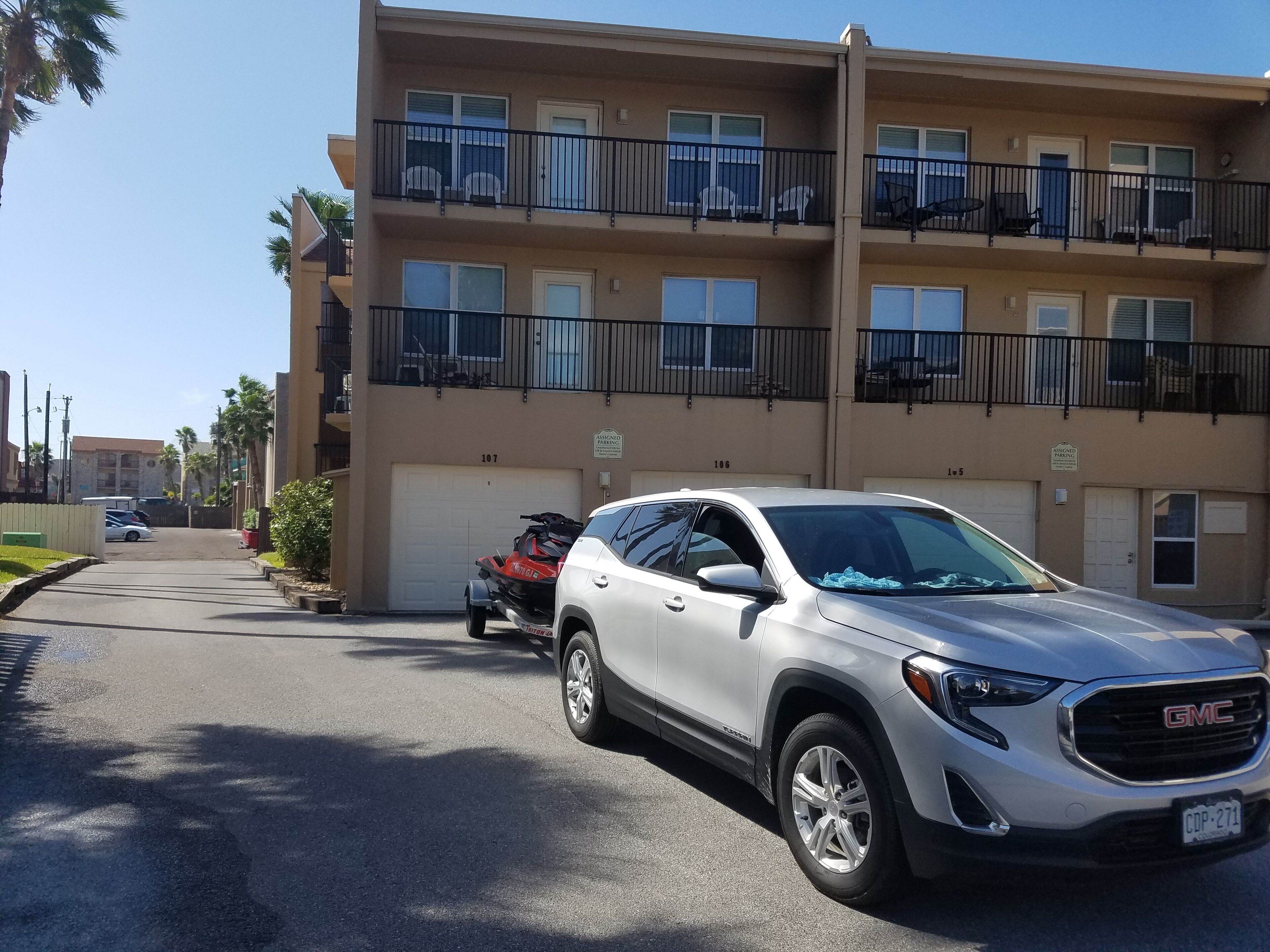 Un edificio residencial de varios pisos con espacios de estacionamiento numerados, una camioneta SUV blanca y un vehículo todo terreno rojo estacionados al frente.