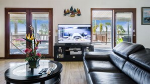 Ein Wohnzimmer mit einer Ledersofa, einem Glastisch mit Fernbedienungen, einem Flachbildfernseher, der ein Rennspiel zeigt, und einem Balkon mit Blick auf Palmen.
