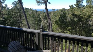 A wooden deck with a bench and a railing overlooking a forested area with a view of the ocean.