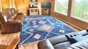 Una sala de estar acogedora con un tapete azul estampado grande, sofás de cuero, vigas de madera en el techo, un televisor de pantalla plana y una ventana con vistas a árboles.