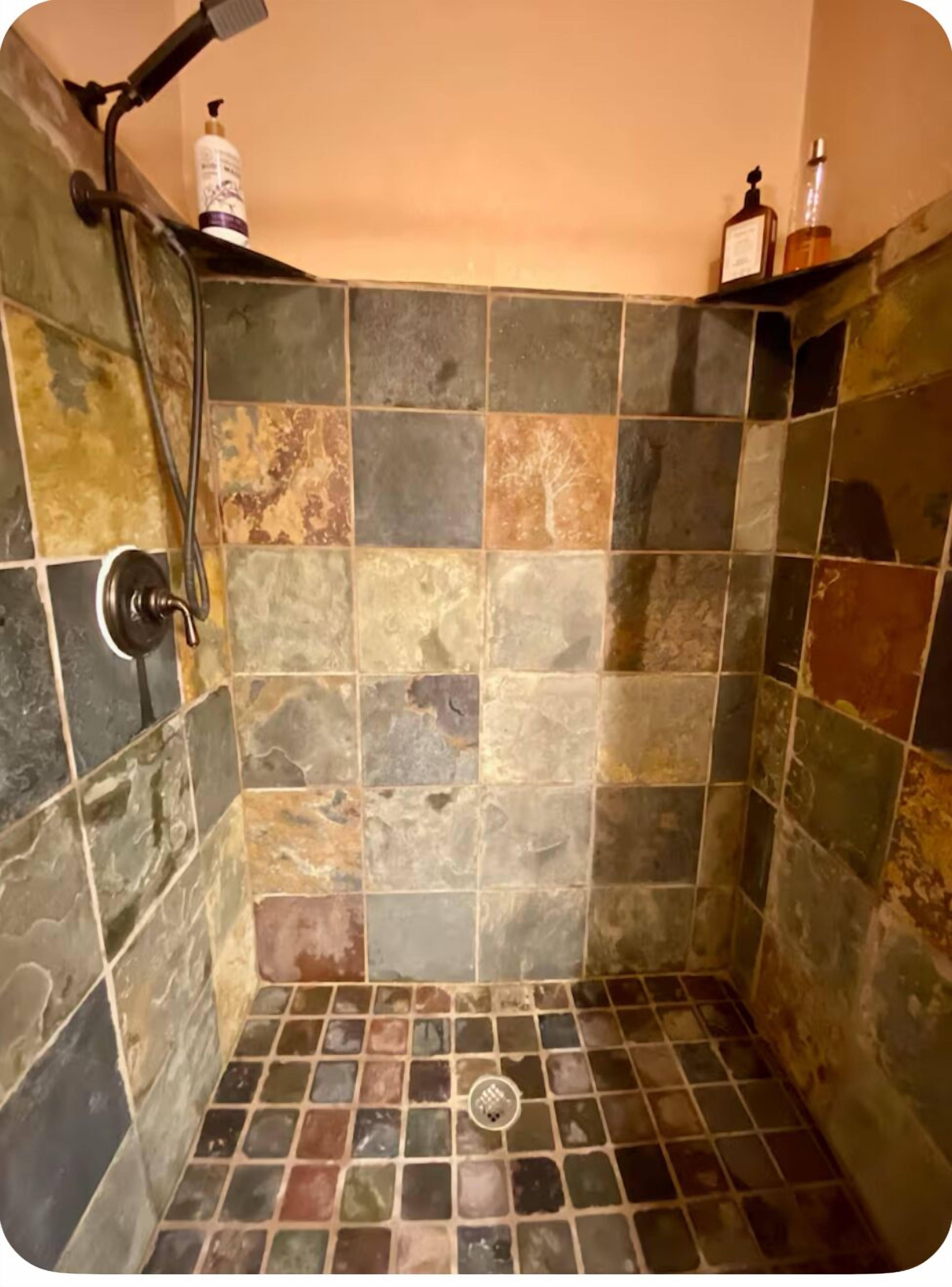 A shower with a tiled wall and floor, a handheld showerhead, and a bottle of shampoo on the wall.