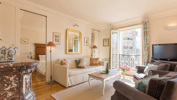 A living room with a sofa, a coffee table, a TV, and a large window with a view of buildings. - Romantic 2 Bed with Peekaboo Eiffel View in Foodie Heaven (Paris)