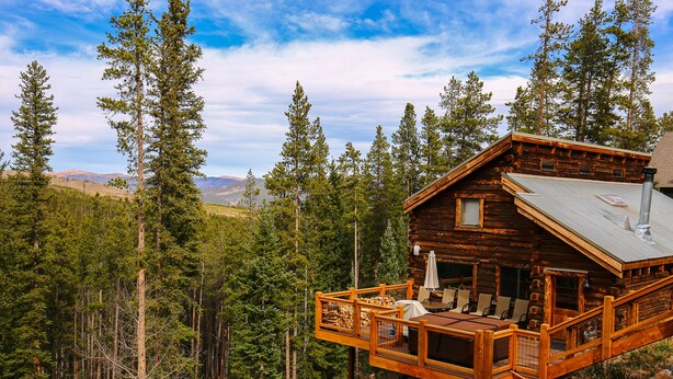 Une cabane en rondins dotée d’une terrasse, entourée de grands pins et avec des montagnes en toile de fond.