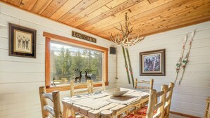 L’intérieur d’une cabane en rondins, avec une table à manger, des chaises, un lustre, des tableaux encadrés et une fenêtre donnant sur une forêt.