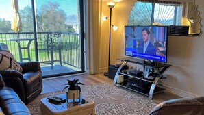 A living room with a leather sofa, a coffee table, a TV, and a balcony with a view of a green lawn. - Remodeled, larger  2 bedroom 2 bath. GOLF/BRIDGE VIEW. WOOD FLOORS BIG SCREEN TV (Lake Havasu City)