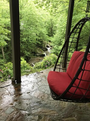 Un fauteuil à bascule rouge avec coussin, placé dans un espace abrité, d’où l’on peut admirer un ruisseau bordé d’arbres.